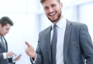 young man with beard in a suit smiling with his hand extended in greeting